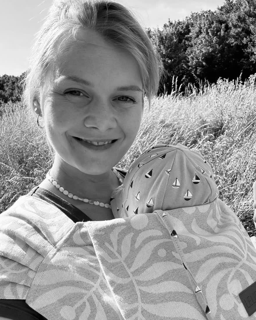 Marie, Dreifachmama und Creatorin, lächelt mit Baby im Tragetuch in der Natur (Schwarz-Weiß-Foto)